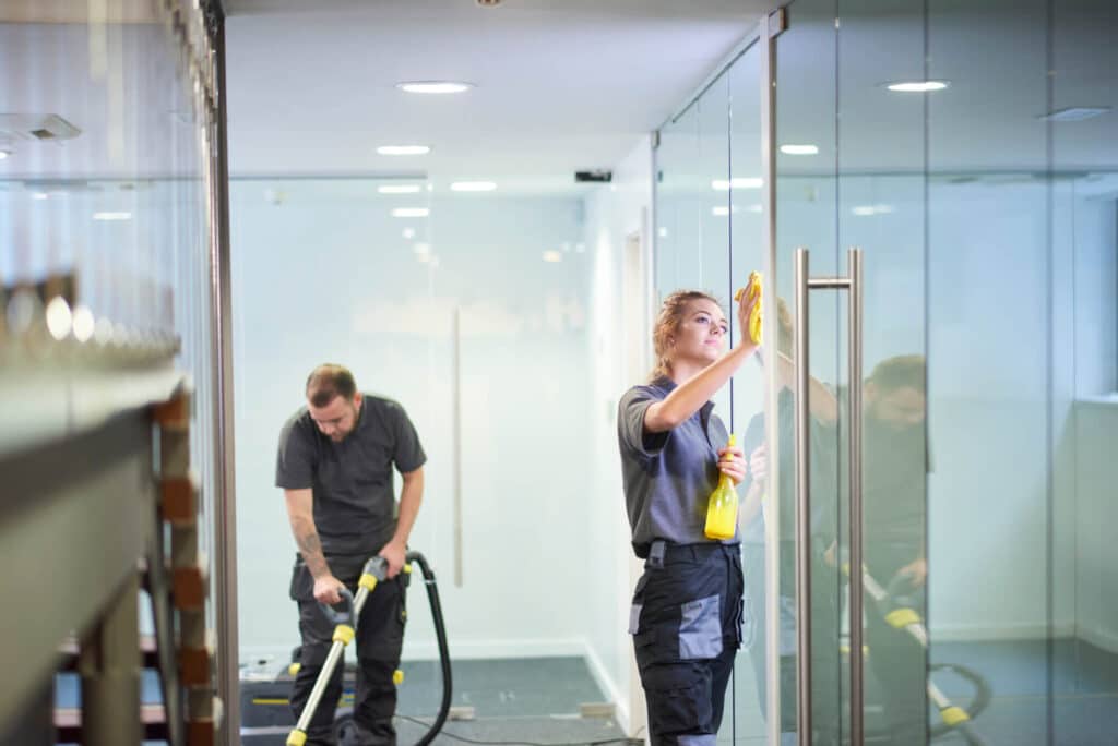 Woman cleans conference room windows while a man is using a floor cleaner to clean carpets