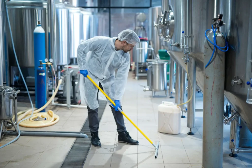 Main in white PPE gown and hat using a floor squeegee in an industrial setting.