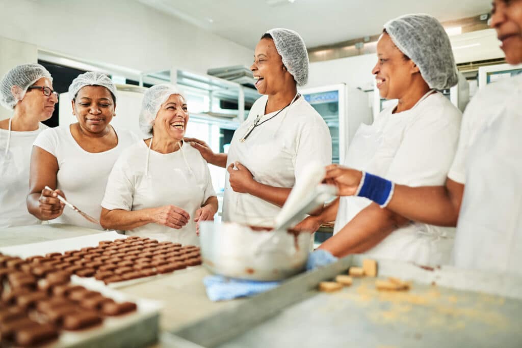 Six women in white clothing and protective hair nets standing around a table laughing while preparing chocolate.