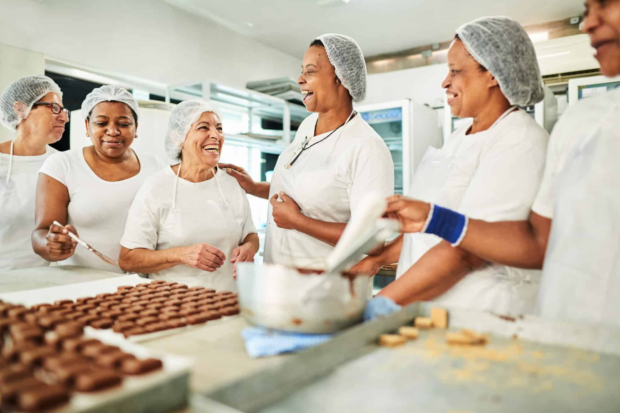 Six women in white clothing and protective hair nets standing around a table laughing while preparing chocolate.