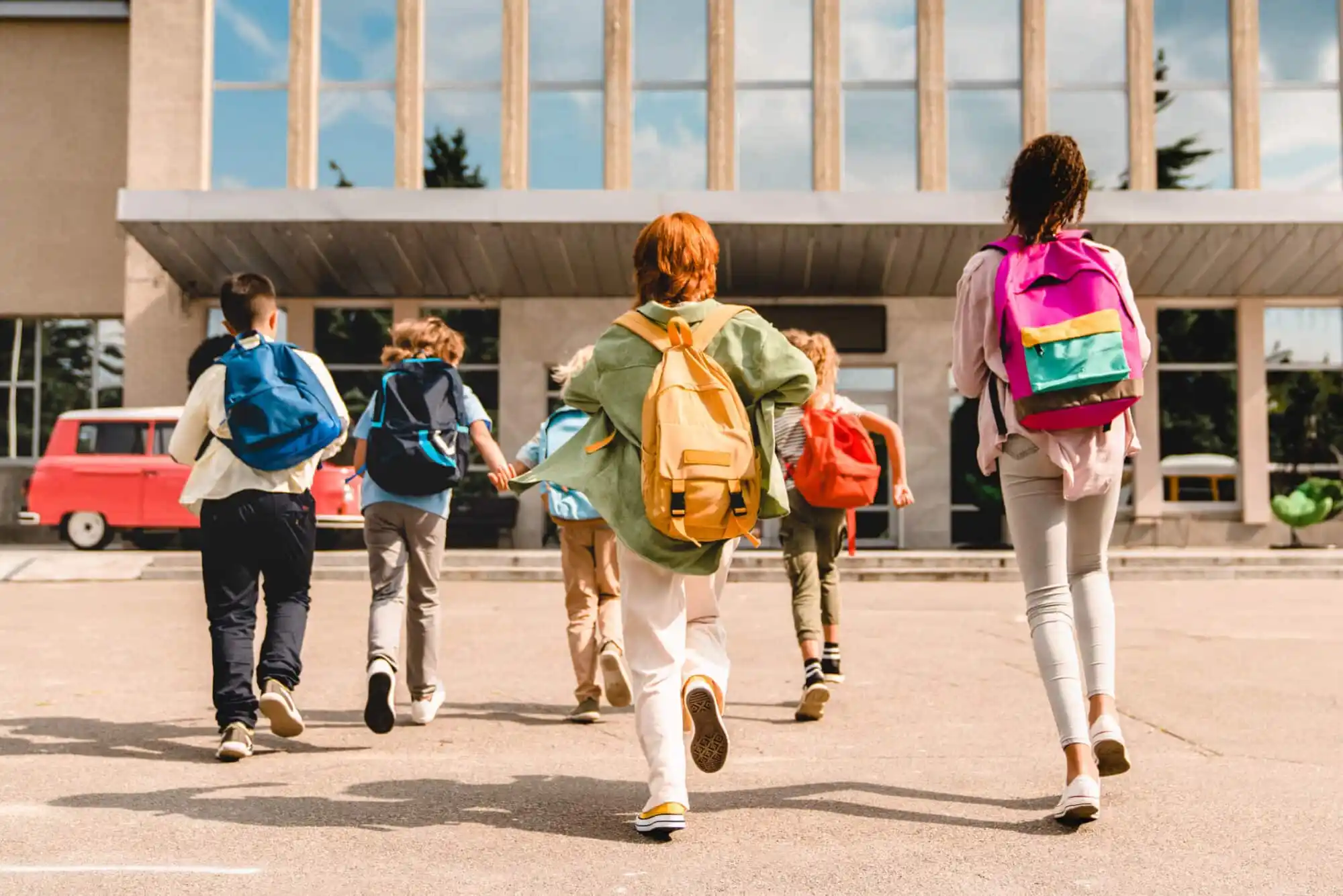 Six children wearing backpacks walking towards a school building.